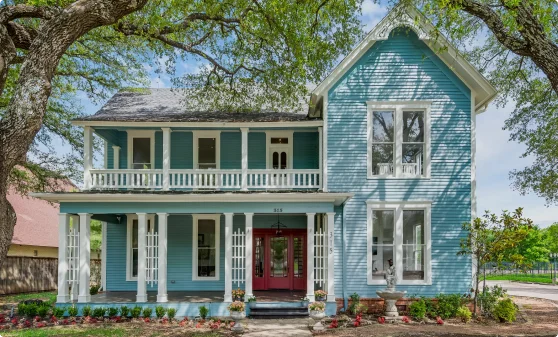 The image depicts a charming two-story house painted in a light blue color with white trim. It features a front porch with decorative columns and railings, large windows, and a red double door entrance. The house is surrounded by mature trees, neatly maintained landscaping, and a stone walkway leading to the entrance.