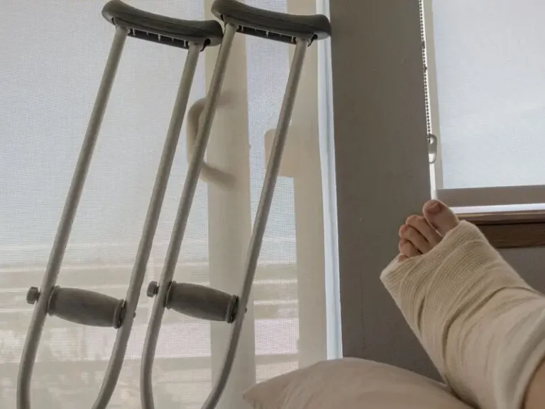 A foot in a cast rests on a pillow near a window. Crutches are leaning against the wall beside it.