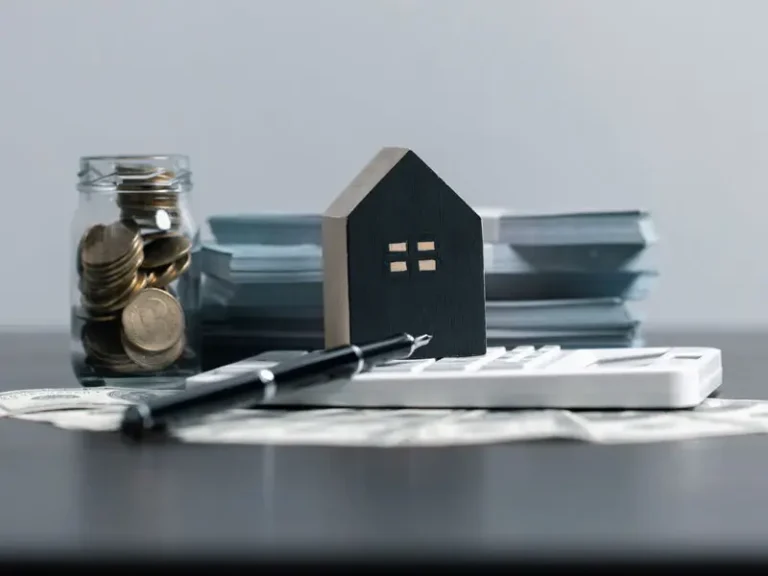 Miniature house model with calculator, pen, and money jar on documents, suggesting financial planning.