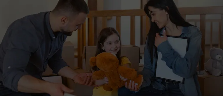 A man, a woman, and a young girl smiling indoors with a teddy bear and framed picture.