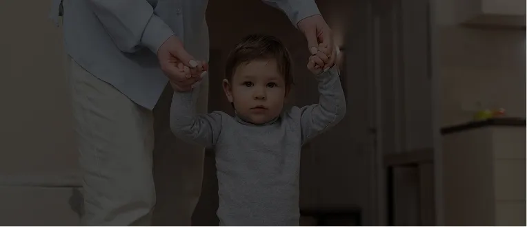 Adult assisting toddler with walking indoors, both wearing casual clothing.