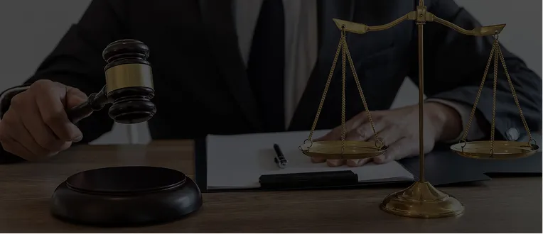 Person in dark suit holding wooden gavel with brass scales of justice on desk.