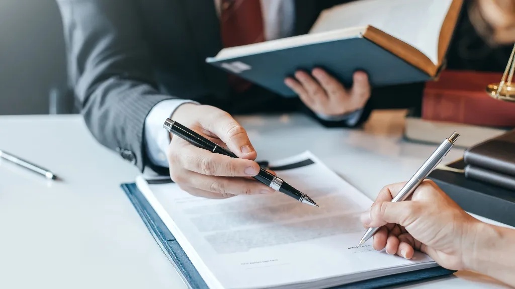 Two professionals in business attire reviewing and signing a document on a white table.
