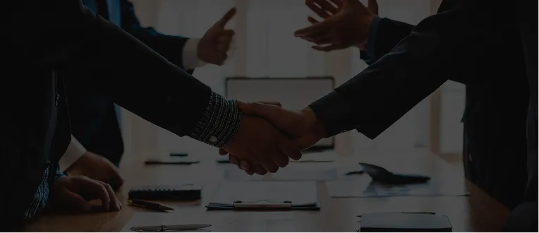 Two individuals in business attire shaking hands over a conference table with documents and a laptop.