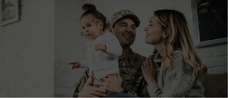 A man in military uniform holding a young child, smiling woman beside them indoors.