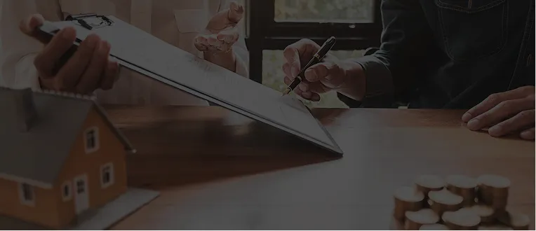 Two individuals at a table reviewing documents, with a pen, clipboard, model house, and stacked coins visible.