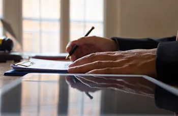 Person writing on paper with pen, reflected on glossy table surface.