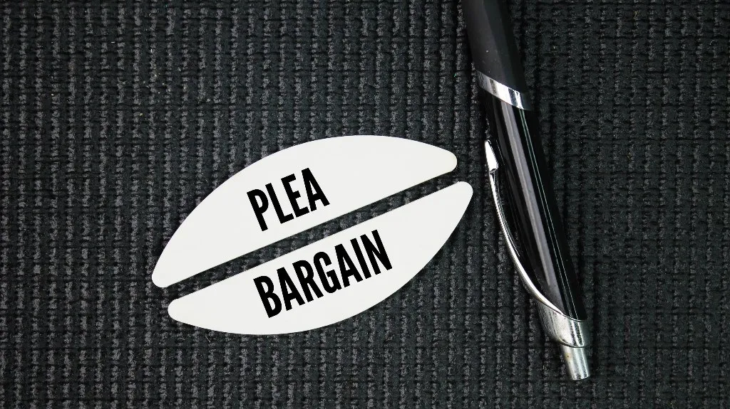 Black pen and white paper cutout with text "PLEA BARGAIN" on dark textured surface.