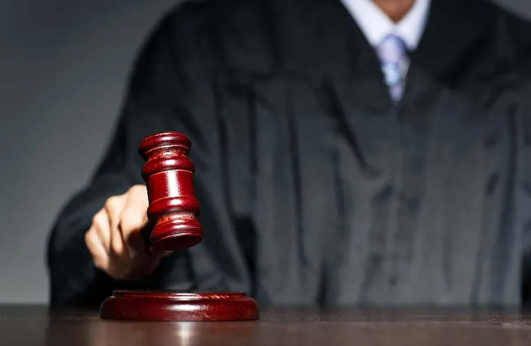 Judge in black robe holding wooden gavel above sounding block on desk.