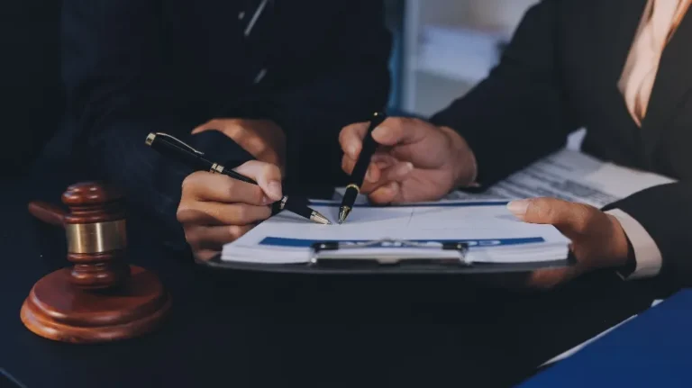 Two professionals in dark suits reviewing and signing documents on a clipboard, wooden gavel nearby.