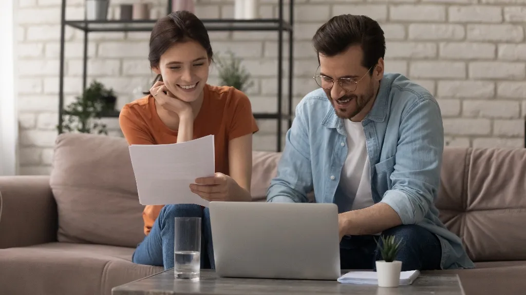 Young woman and man smiling while reviewing documents and using laptop on sofa.