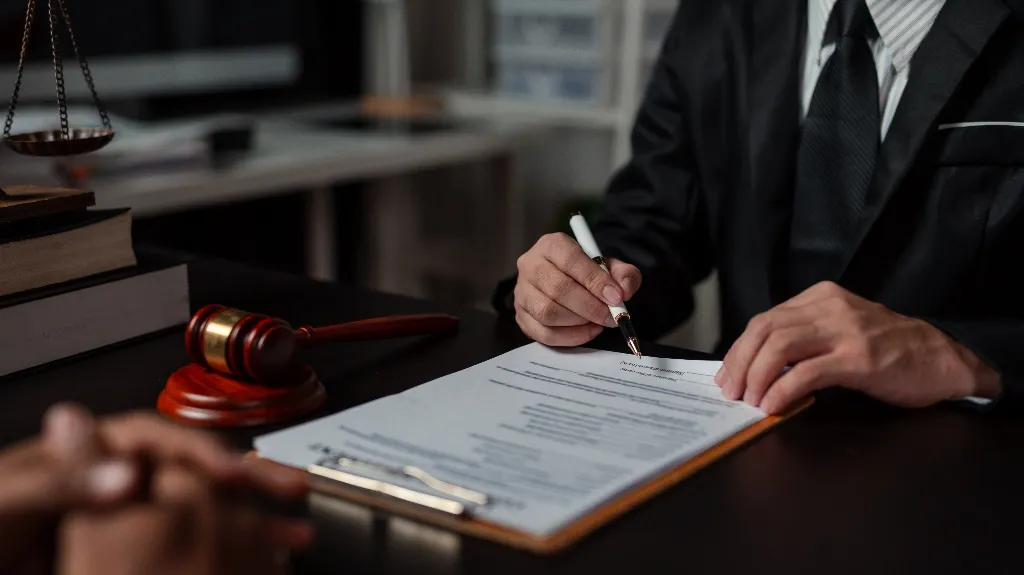 Person in formal suit signing legal document on clipboard with gavel and law books nearby.