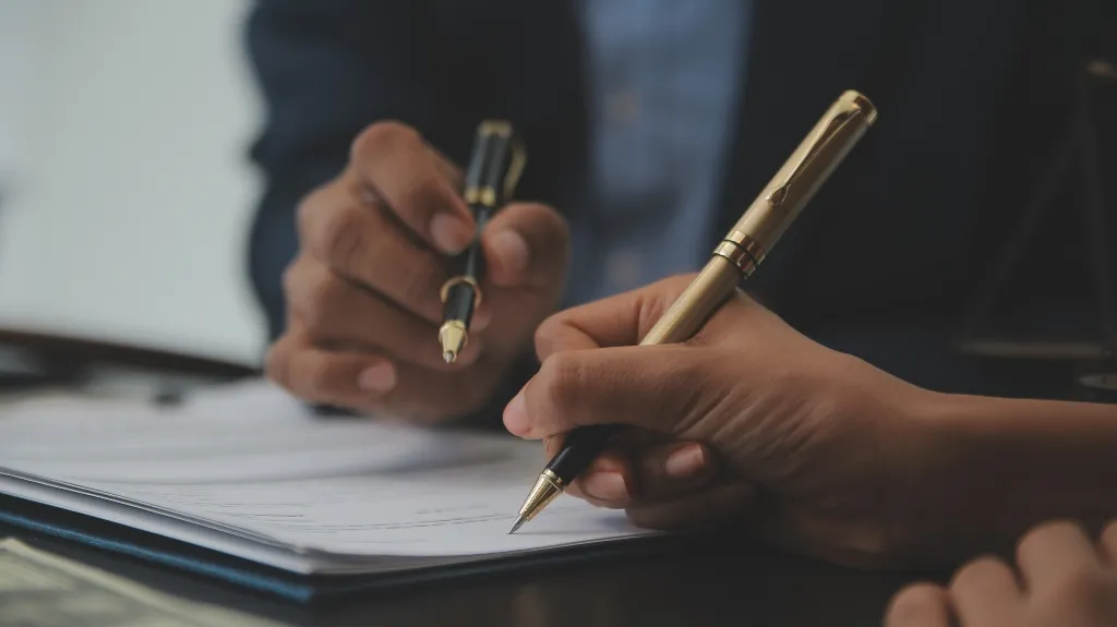 Two hands holding gold and black pens poised over a document on a desk.