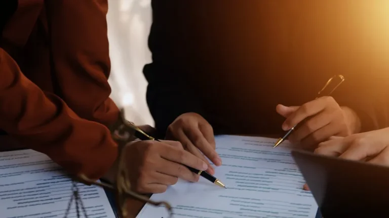 Two individuals reviewing and annotating documents with pens under warm lighting.