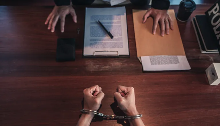 Person in handcuffs facing a desk with documents and police folder, hands visible.