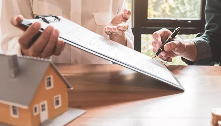 Two individuals reviewing and signing a document on a clipboard, with a small house model on the table.