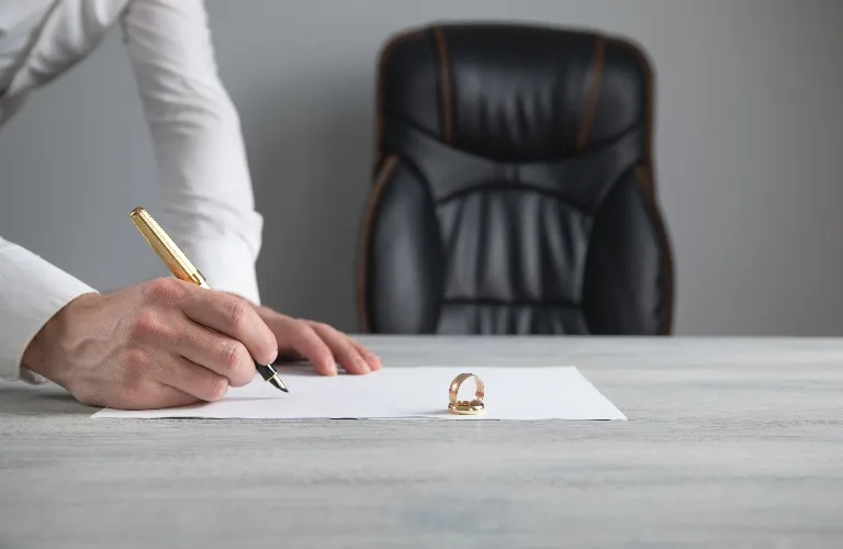Person signing a document on a desk with a gold wedding ring placed on the paper.