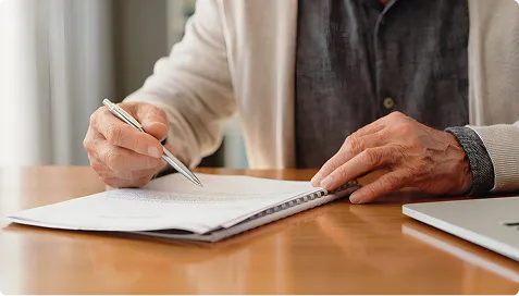 Person in beige sweater writing on a spiral-bound notebook with a silver pen at a wooden desk.