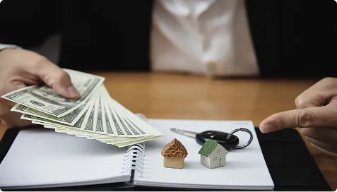 Person holding US dollar bills and pointing at small house models and car keys on notebook.