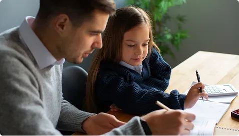 Adult male assisting young girl with homework at wooden table with calculator.