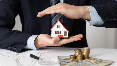 Person in suit holding a small model house above a desk with money and financial documents.