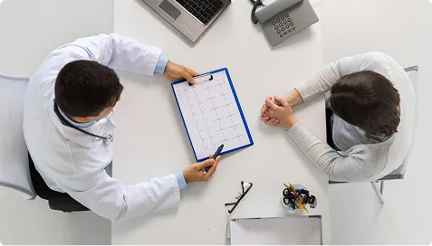 Overhead view of doctor in white coat discussing calendar with patient at desk.