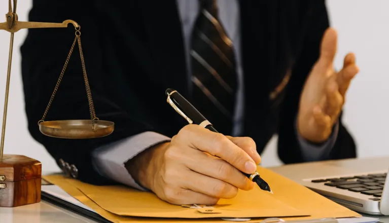 Professional in suit writing on a folder with a pen, scales of justice beside.