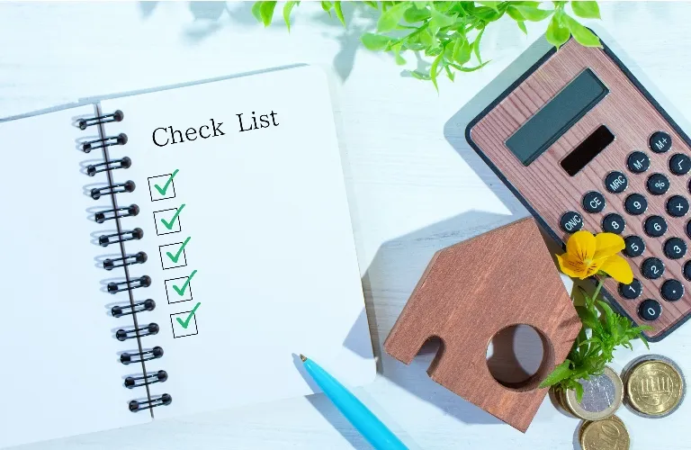 Open checklist notebook with all items checked, blue pen, wooden house model, calculator, coins, and green plant.