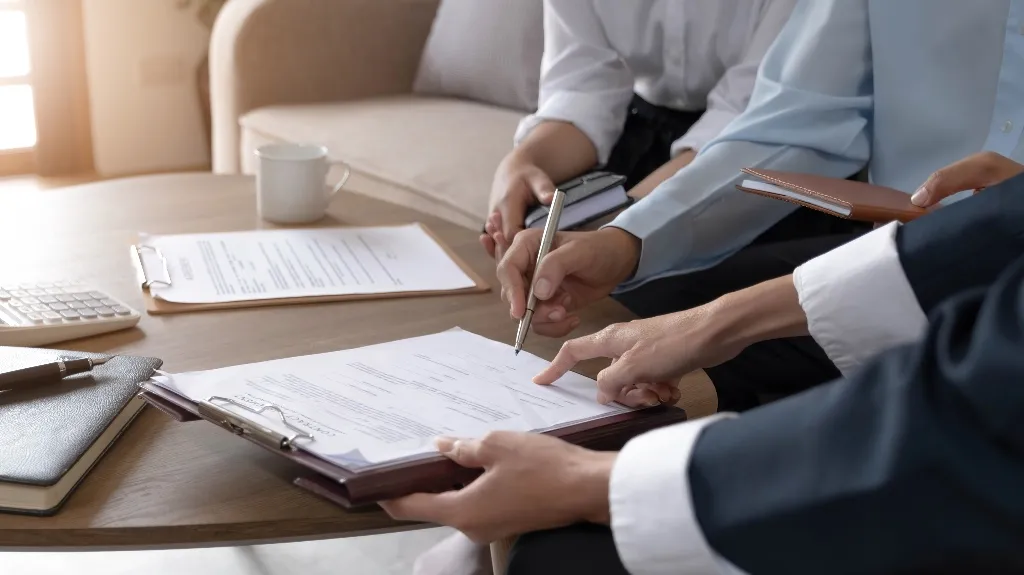 Three professionals reviewing and signing documents at a wooden table with notebooks and a coffee cup.