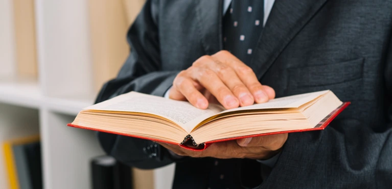 Person in dark suit and tie holding and reading an open book with a red cover.