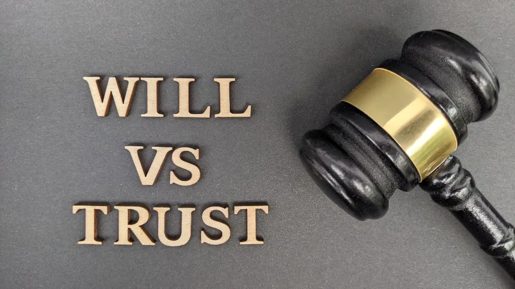 Wooden letters spelling "WILL VS TRUST" beside a black and gold judge's gavel on gray background.