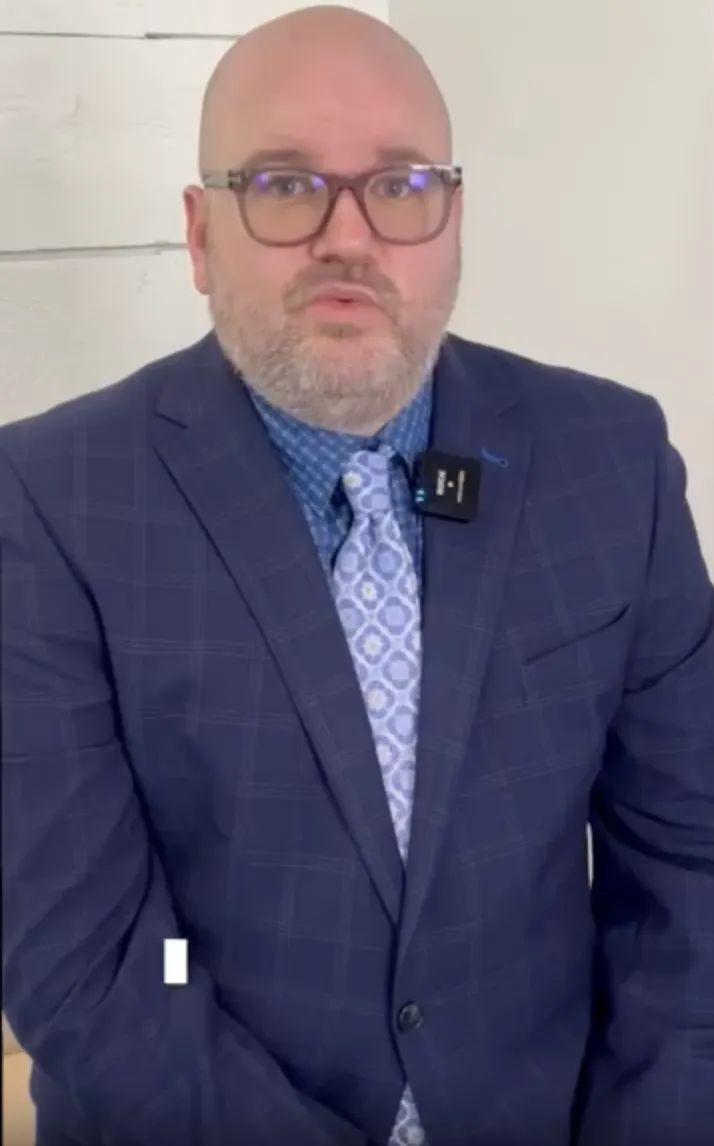 Professional middle-aged man wearing glasses, a blue checkered suit, patterned tie, and a lapel microphone.