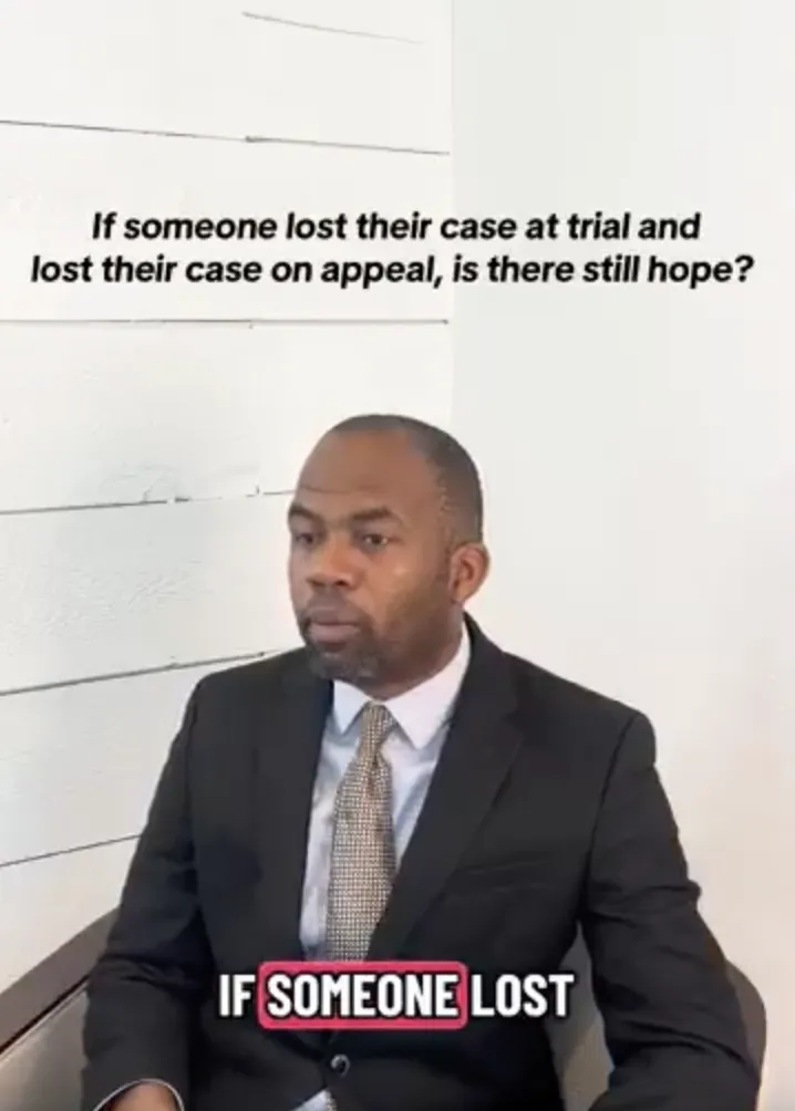 Man in a dark suit and tie seated against a white wall with legal text above him.