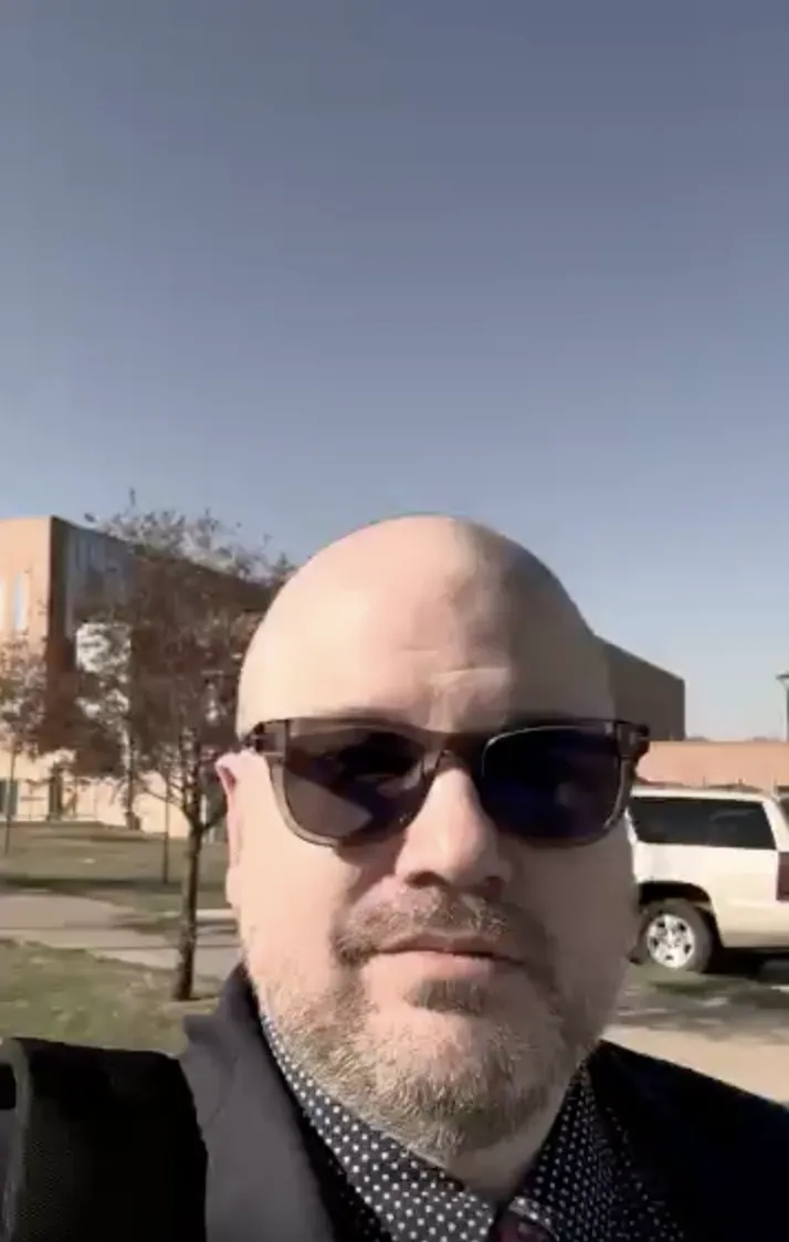 Middle-aged man with sunglasses and a beard outdoors, buildings and parked cars in background.