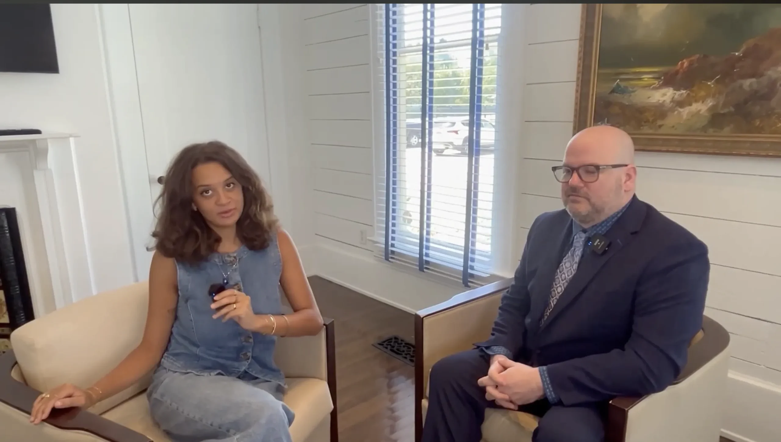 Two individuals seated in a well-lit room with white walls, window blinds, and a framed painting.