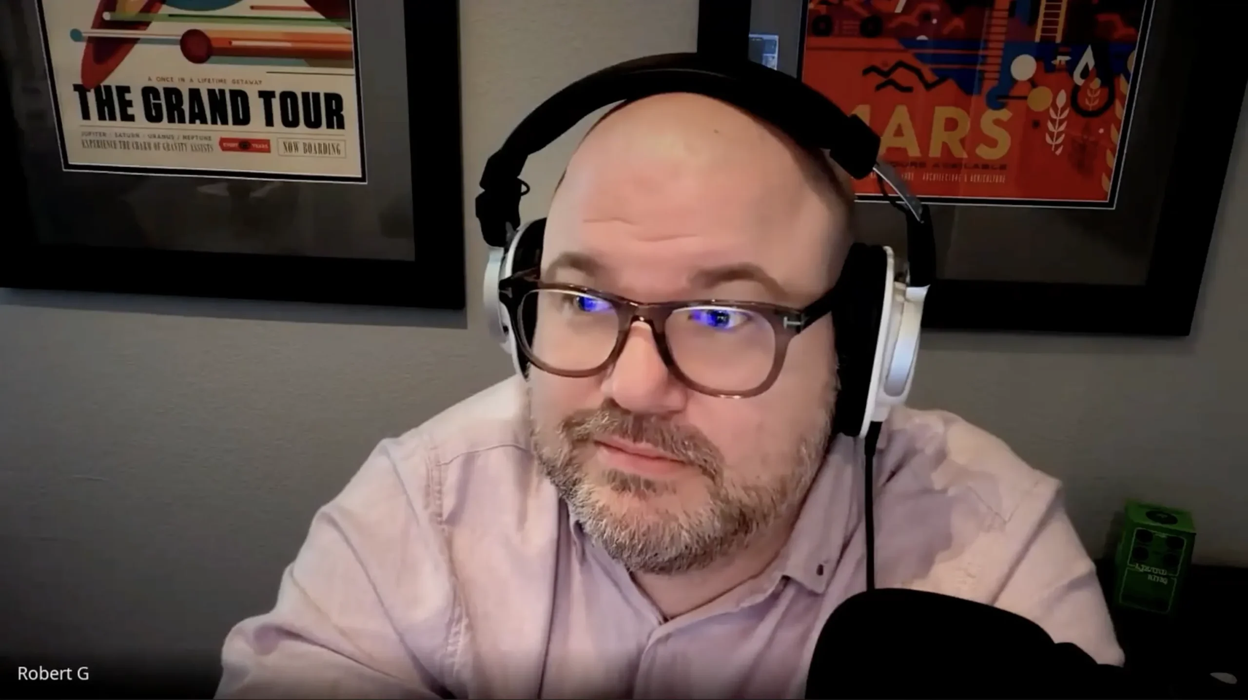 Middle-aged man with glasses and headset, seated indoors with framed posters behind.