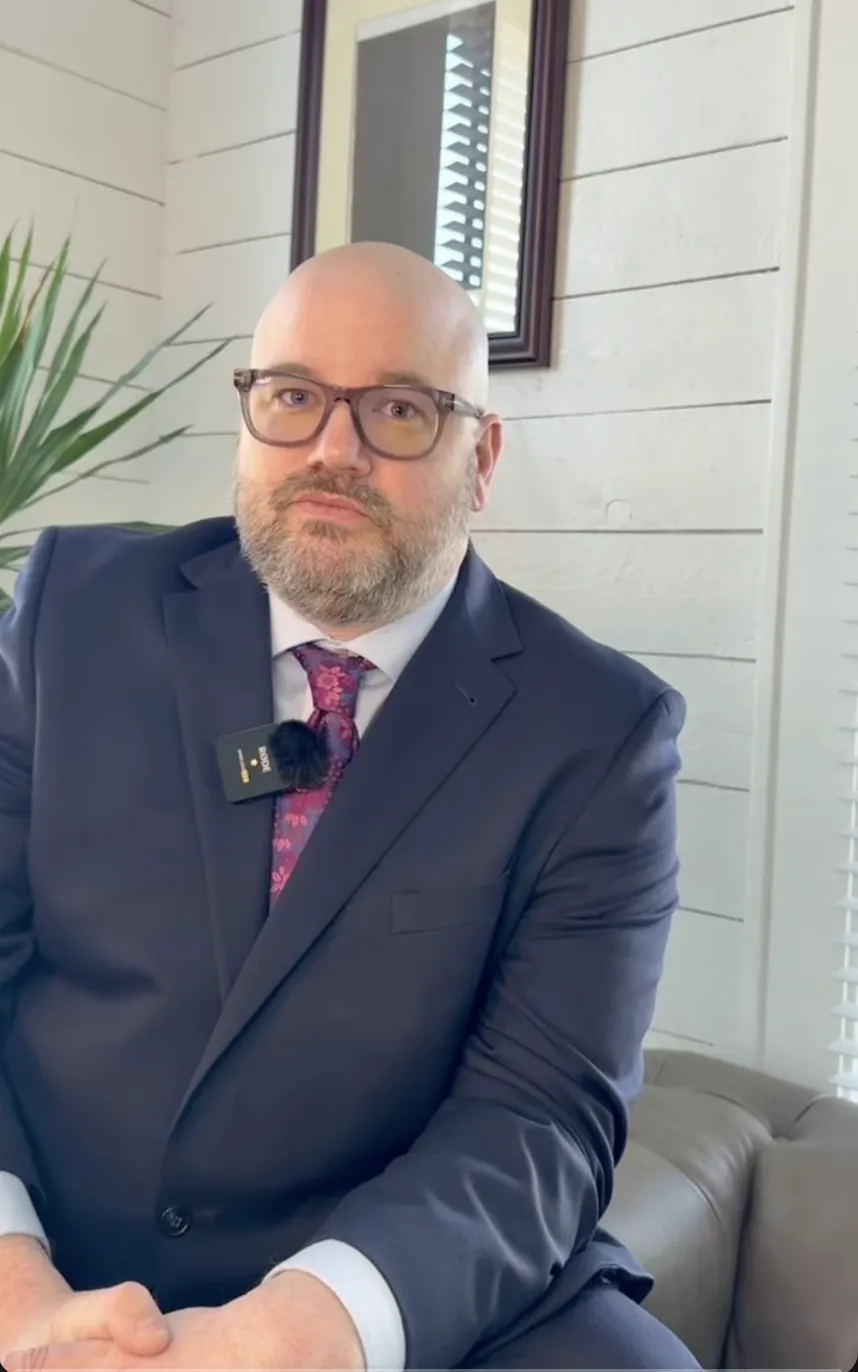 Professional bald man with glasses wearing a navy suit and purple tie seated indoors.