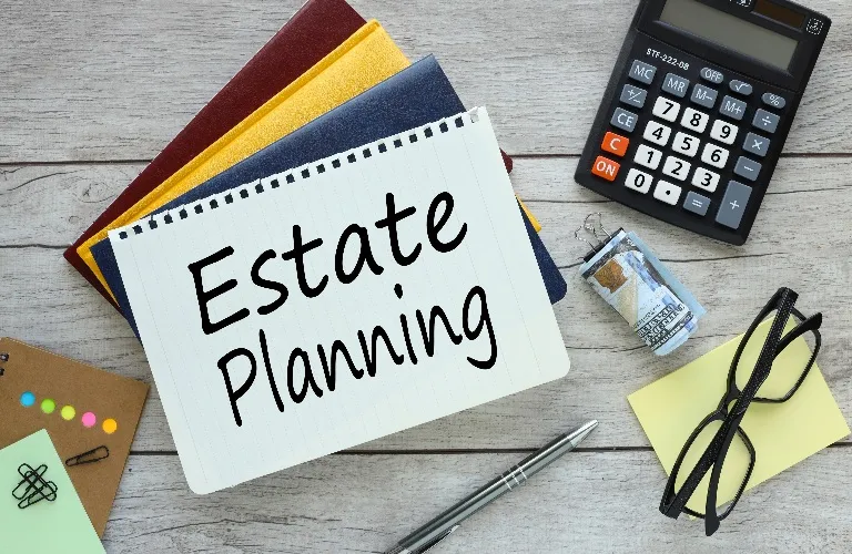 Notepad with "Estate Planning" text, calculator, pen, eyeglasses, and folders on wooden surface.