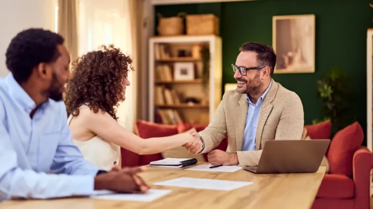 Three professionals in business attire shaking hands across a wooden table with a laptop and documents.