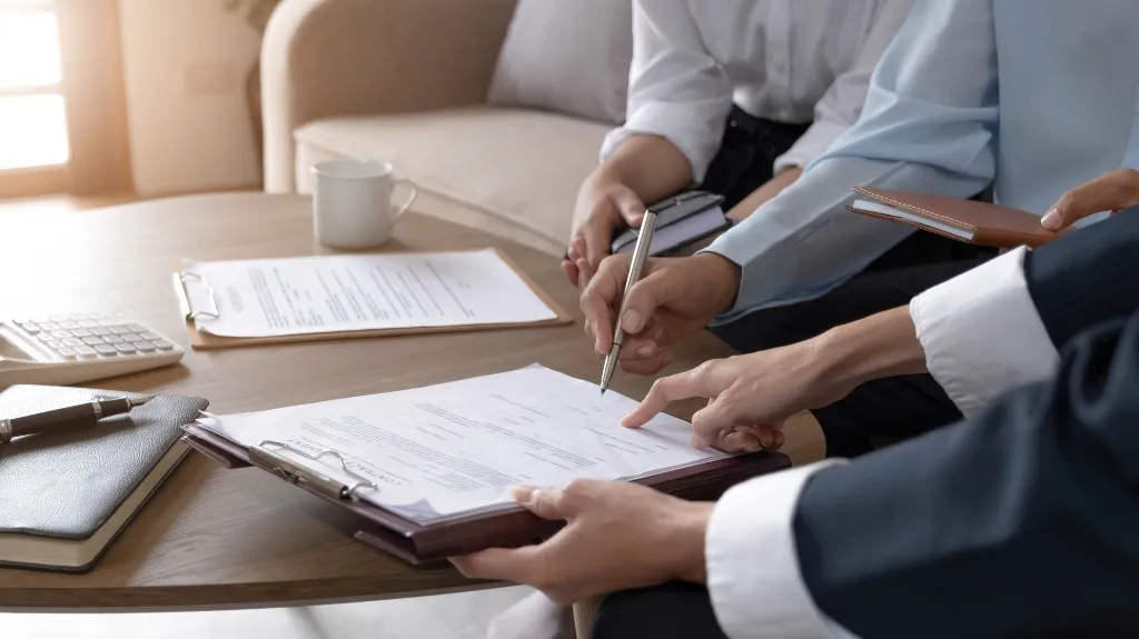 Three professionals reviewing and signing documents on a wooden table with office supplies.