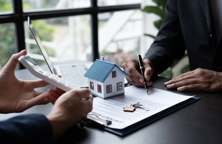 Two professionals reviewing a contract with a house model, keys, and a laptop.