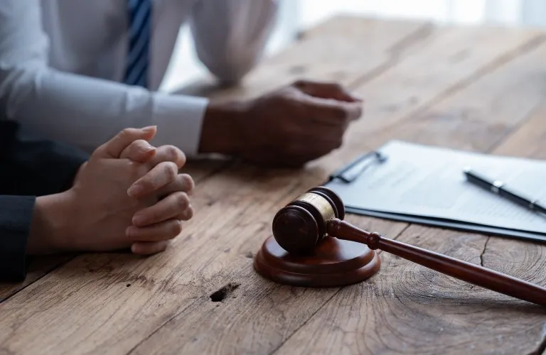 Wooden judge's gavel on table with clasped hands and legal documents in background.