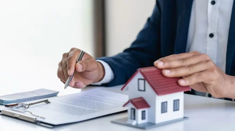 Person in business attire holding a pen and touching a small house model beside a contract and keys.