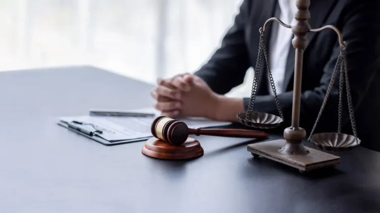Wooden gavel, brass scales of justice, and folded hands of a person in formal attire on desk.