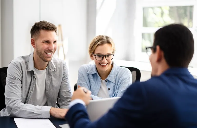 Three professionals in a bright office engaged in a smiling discussion around a laptop.