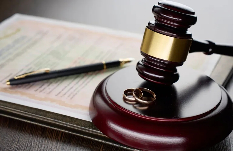 Wooden judge's gavel with two gold wedding rings on base, pen and legal document in background.