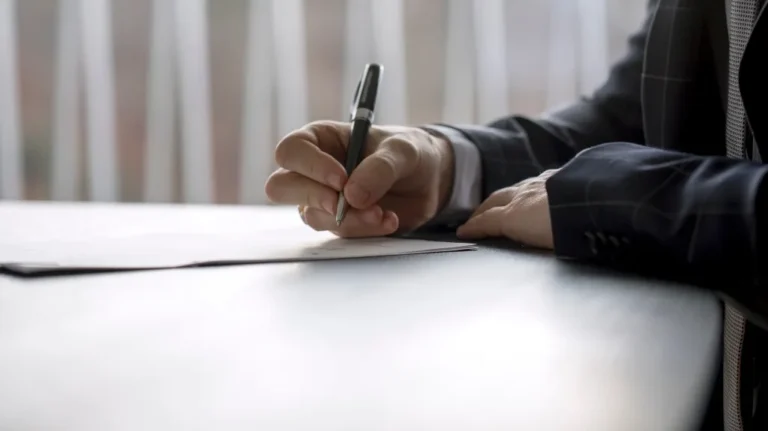 Close-up of a person in a dark suit writing with a pen on paper at a desk.