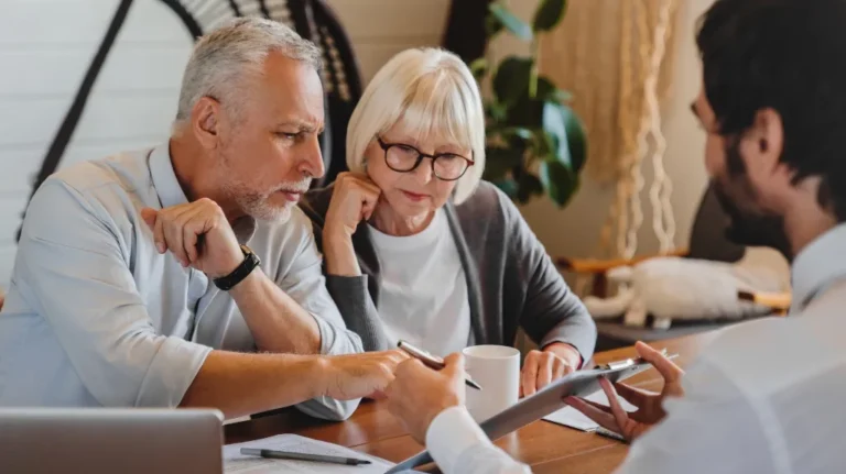 Three adults engaged in a discussion, reviewing documents at a wooden table indoors.