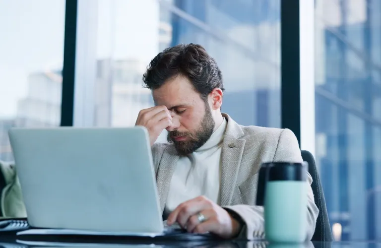 Man in beige blazer sitting at desk with laptop, rubbing eyes in modern office.