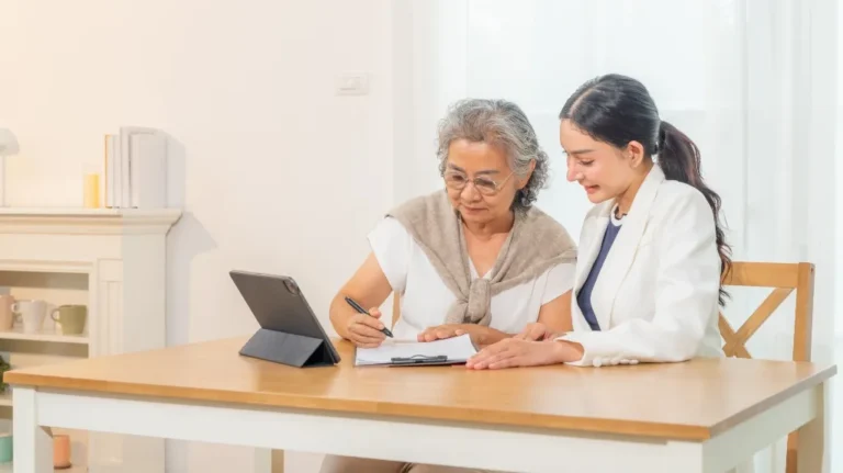Two women, one elderly and one younger, seated at a wooden table with a tablet, reviewing documents together.